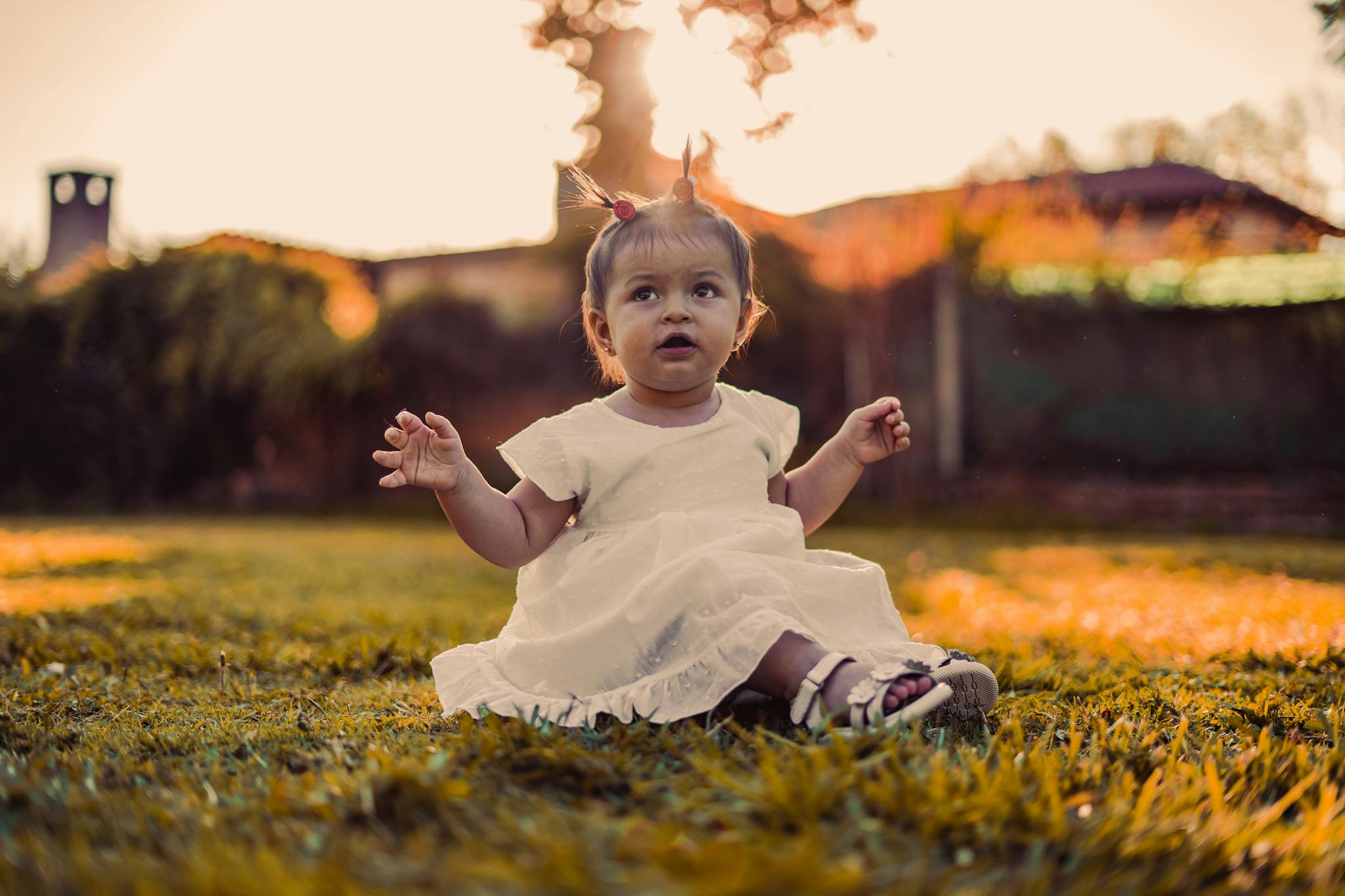 A cute baby girl in a white dress plays on grass during sunset in a park setting.
