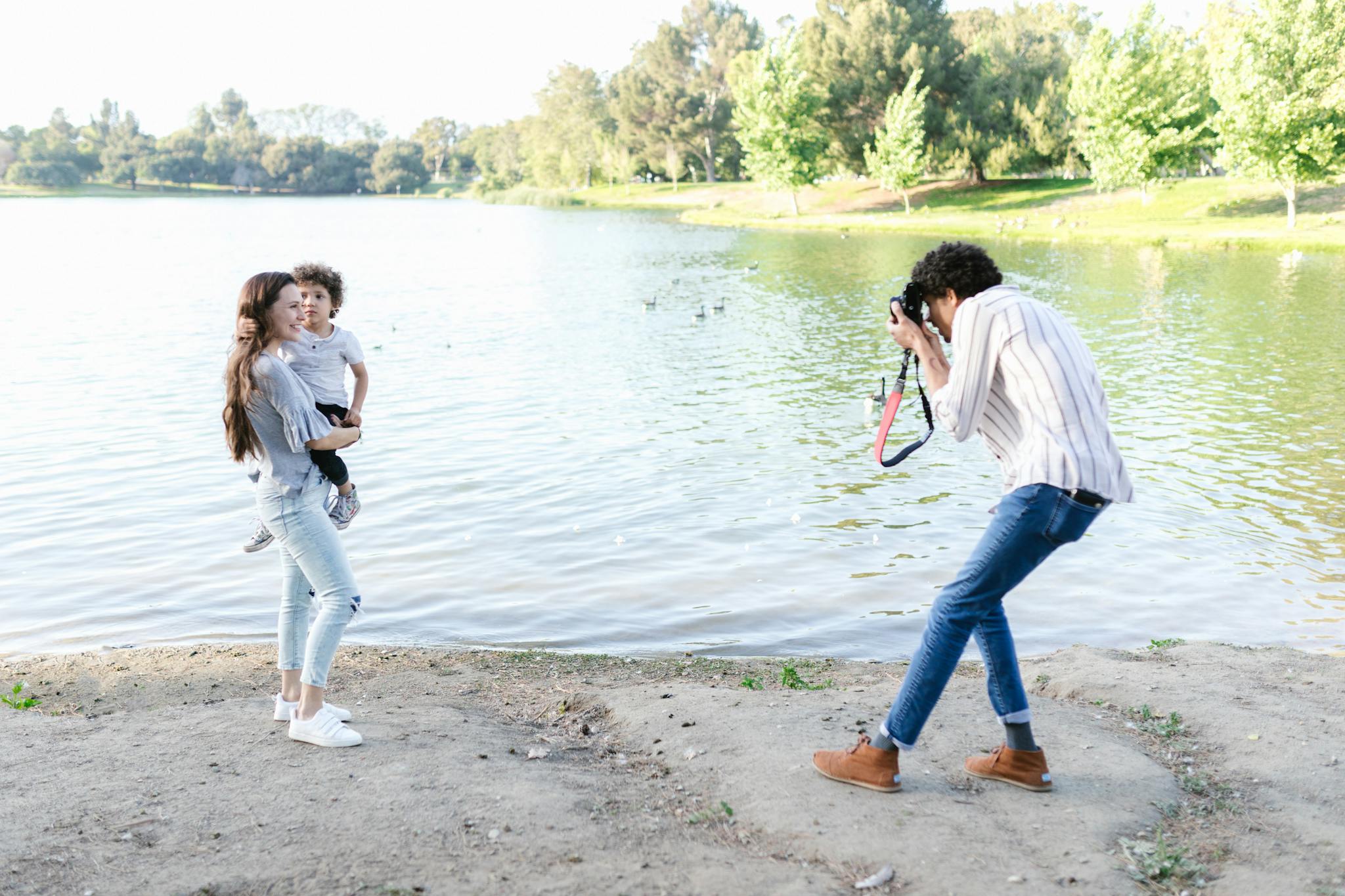 A photographer captures a mother and child by a serene lakeside in a lush park.
