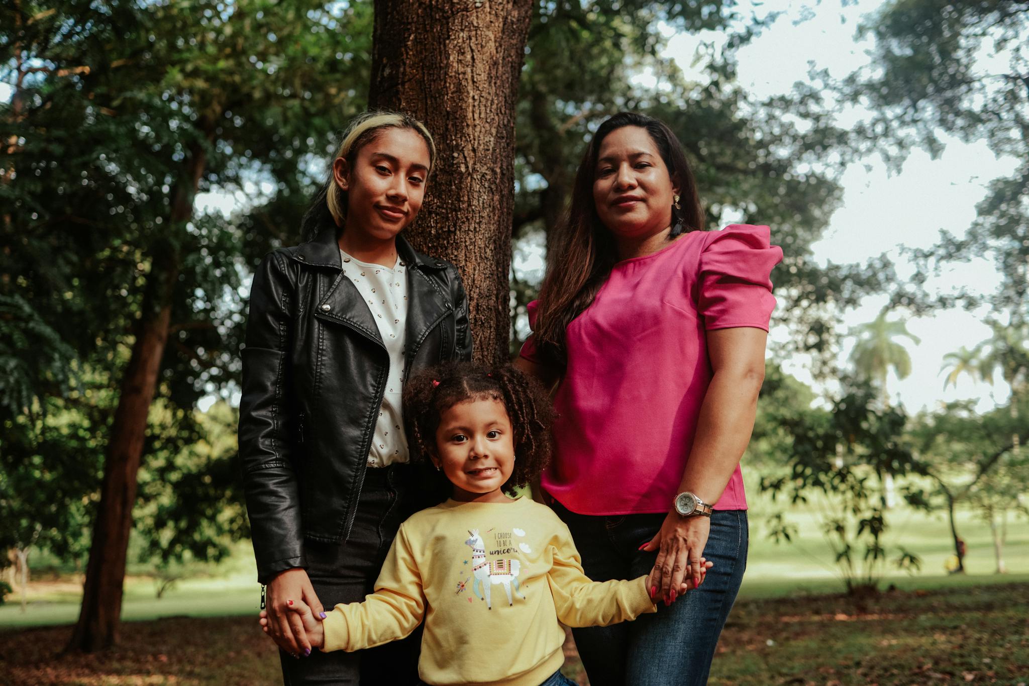 Family trio enjoying a day in the park under the trees, capturing a candid moment of togetherness.